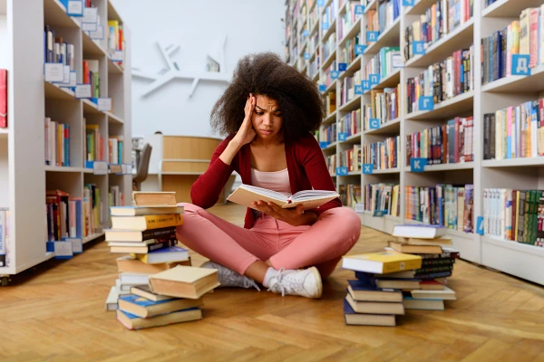 A woman sits in a library surrounded by stacks of books, looking frustrated while reading.